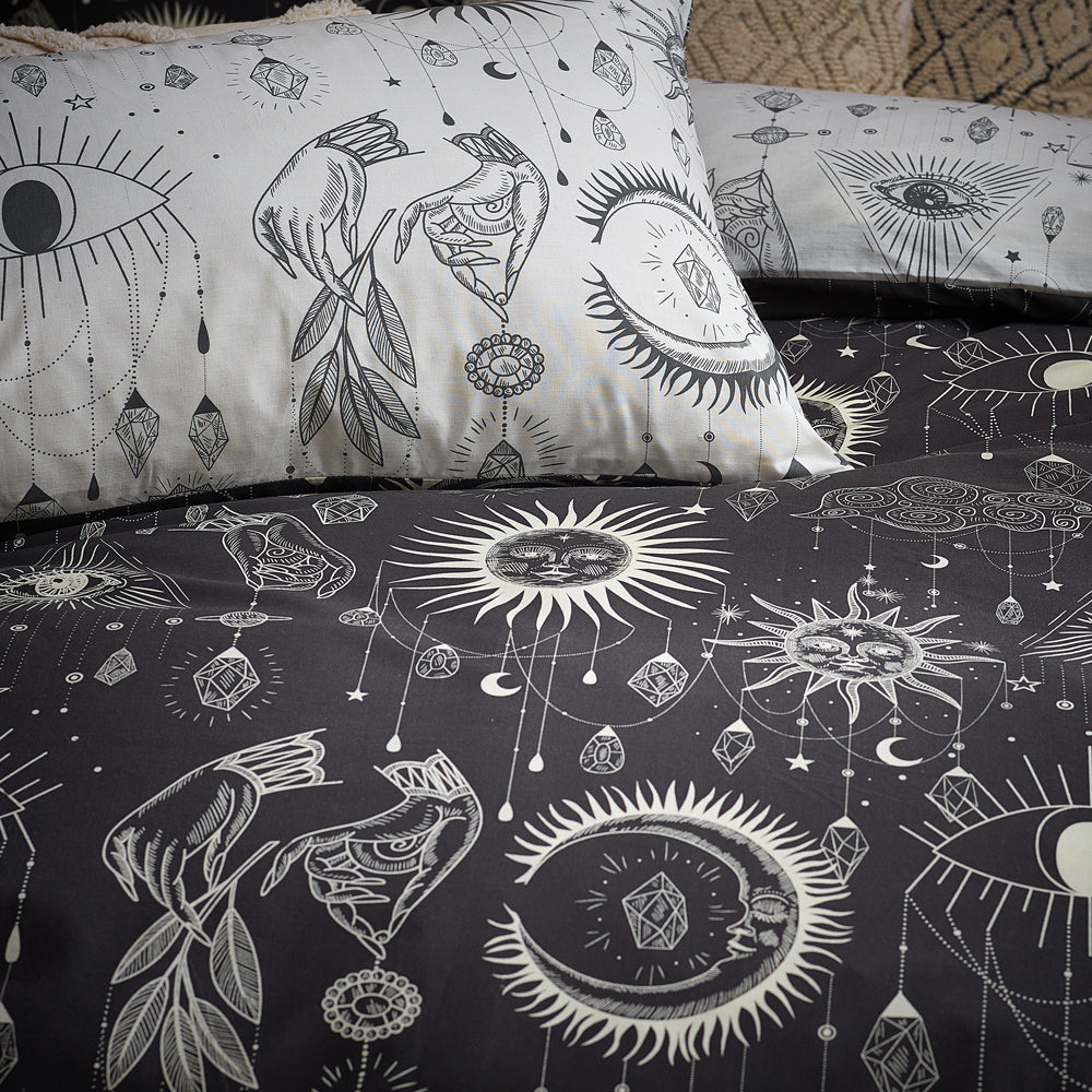 Patterned bedding with celestial and mystical motifs, such as suns, moons, eyes, and birds, in white on dark and light backgrounds, arranged on a bed.