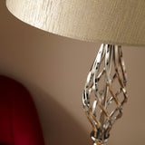 A textured lampshade casts light on a twisted metallic lamp base, with a soft-focus red object in the background.