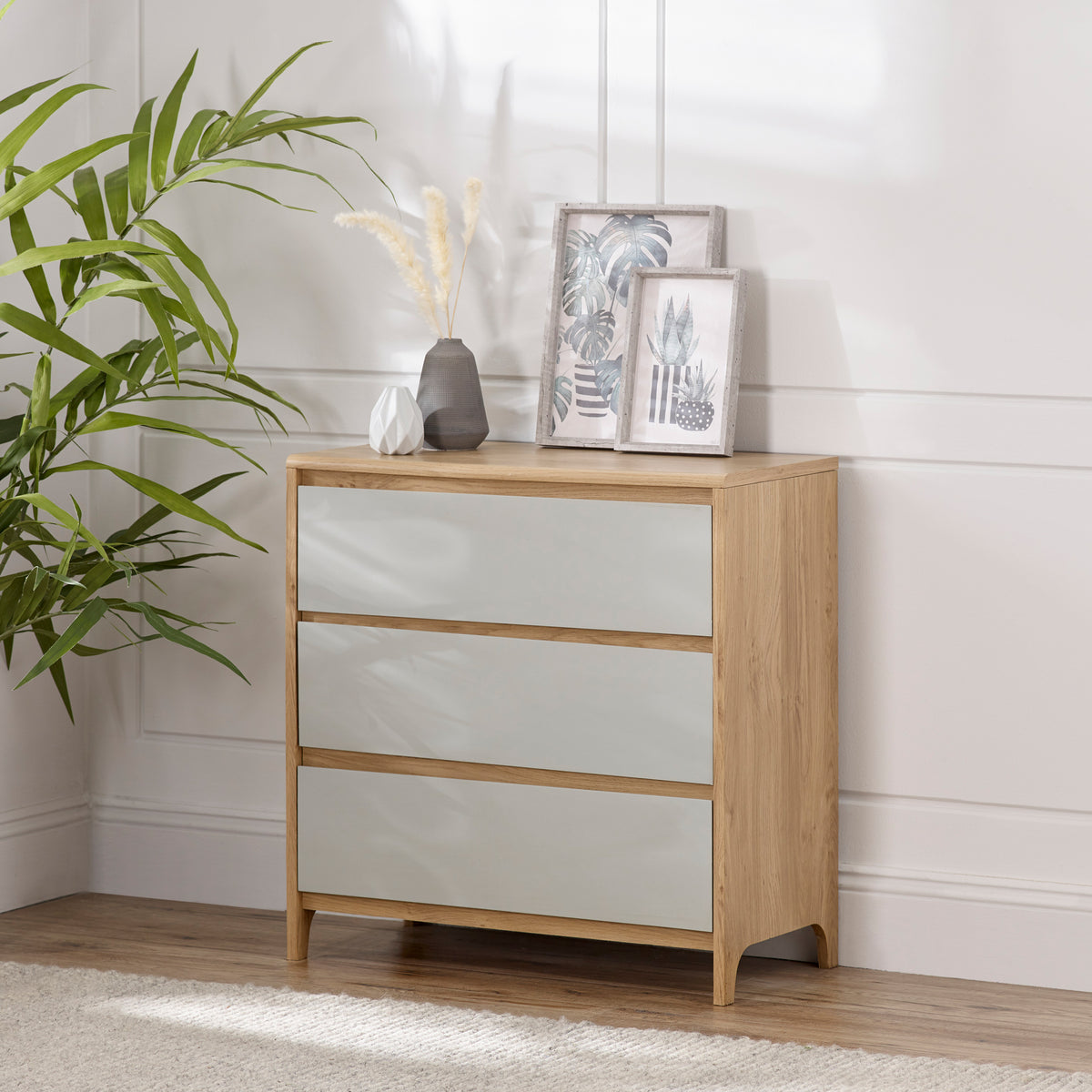 A wooden dresser with frosted glass drawers stands against a wall, adorned with a vase, decorative geometric object, and framed pictures, beside an indoor plant.