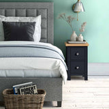 A neatly made bed with a grey headboard is centered, adjacent to a nightstand with decorative items. The room has a soft green wall and wooden flooring.