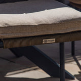 A close-up of a chair cushion with textured fabric on a braided outdoor chair; a logo "maze" visible.