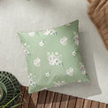 A green floral-patterned pillow rests on a wooden floor beside a beige throw and a decorative green item, partially on a white woven mat.