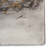 A textured fabric with gray and gold patterns, likely a corner of a rug, set against a plain backdrop.