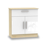 A small wooden cabinet with white doors and a drawer, featuring metal handles, stands isolated against a plain white background.