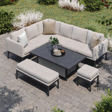 Beige sectional outdoor sofa arranged in an L-shape around a dark slate coffee table on a paved patio, with surrounding greenery. Two ottomans complement the setting.