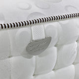 A close-up of a textured white mattress featuring a circular pattern with a metal tag reading "Spencer MATRAH" attached by a striped cord.