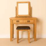 A wooden vanity table with two drawers and a square mirror, accompanied by a cushioned stool, set against a neutral wall and carpeted floor.