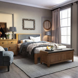 A neatly made bed with gray and brown bedding is centered in a well-lit bedroom, adorned with wooden furniture, decorative mirrors, and framed artwork.