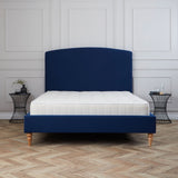 A blue upholstered bed with a white mattress stands stationary against a white paneled wall, with a black side table nearby, set on a herringbone wood floor.