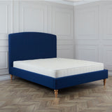 A blue upholstered bed with a white mattress stands against a white paneled wall on a herringbone-patterned wooden floor.