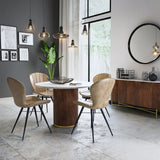 A modern dining room with a round marble table, four tan chairs, and hanging pendant lights. Artwork, a mirror, and a plant decorate the space.