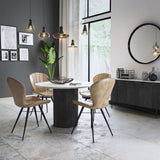 Modern dining area with a circular white table, tan chairs, pendant lights, beside a large window, and a dark cabinet adorned with decor.