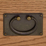 A metal drawer pull mounted on a wooden surface, with a semi-circular handle that pivots at two points.