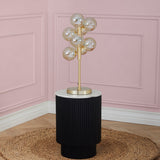 A gold-colored modern table lamp with multiple spherical bulbs stands on a black cylindrical table against a pink wall with paneling.