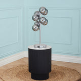 A modern metallic lamp with spherical shades stands atop a ribbed black cylindrical table against a light blue wall and a woven circular mat on the floor.