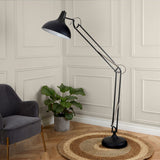 A large black floor lamp with an adjustable arm stands lit in a room, set against a paneled wall, with a plant, chair, and circular rug nearby.