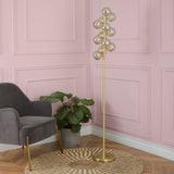 A gold floor lamp with multiple spherical bulbs stands next to a grey armchair and a potted plant, against a pink paneled wall on a wood floor.