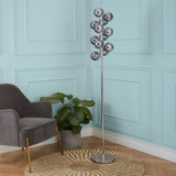 A modern chrome floor lamp with multiple spherical fixtures stands in a room with a grey armchair, a green potted plant, and light blue paneled walls.