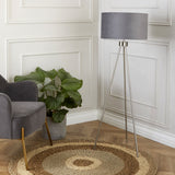 A tripod floor lamp with a gray shade stands illuminated next to a gray upholstered armchair; a large potted plant sits nearby in an elegant paneled room.