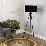 A tall, black tripod floor lamp stands illuminated in a classy room, next to a grey armchair and a potted plant, over a circular woven rug on a wooden floor.