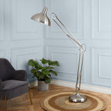 An adjustable silver floor lamp extends over a grey armchair, set against a wall with rectangular moldings. A plant and round rug complete the cozy room corner.