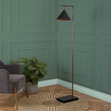 A black-shaded floor lamp with a brass stand illuminates a corner beside a grey armchair, with a green potted plant on a round woven rug against a teal wall.