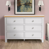 A white six-drawer wooden dresser sits against a pink wall, flanked by black wall lamps, under a framed painting of a Venetian canal scene.