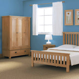 A wooden bed and wardrobe furnish a bedroom with blue walls, white curtains on a window, and a lamp on a bedside table.