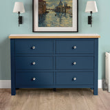 A blue six-drawer dresser stands against a wall with a painting above and flanked by two wall-mounted lamps, in a room with teal walls and wooden flooring.