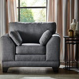 A charcoal gray armchair with two matching pillows is positioned near a round side table with decorative items, against a window with sheer curtains and a tree outside.