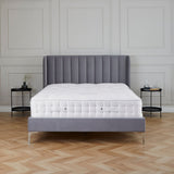 A neatly made bed with a tufted mattress is centered in an elegant room with herringbone wood flooring and sophisticated panelled walls. Two small bedside tables flank the bed.