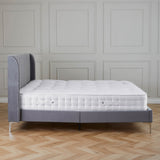 A gray, tufted single bed with a white mattress sits idle in a room with paneled walls and herringbone wood flooring.