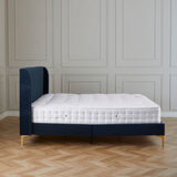 A single bed with a blue upholstered headboard and a white mattress, stands idle against a paneled wall in a room with herringbone wood flooring.