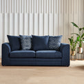 A dark blue sofa with patterned cushions sits against a wall with vertical blinds, accompanied by a plant in a metallic pot to the right.