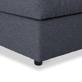 A gray upholstered ottoman with a cushion on top, featuring black rounded feet, sitting against an isolated white background.