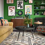 A vibrant living room featuring a green wall adorned with an assortment of framed artworks, flanking a yellow velvet couch and a dark round coffee table over a patterned rug.