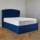 A blue, tufted headboard attached to a matching bed base with a white, quilted mattress in an elegant room with paneled walls and herringbone-patterned wooden floor.