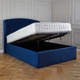 A blue upholstered storage bed with its mattress lifted to reveal an empty compartment underneath. It's situated in a room with paneled walls and herringbone wood flooring.