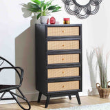 A black, wooden five-drawer dresser with wicker fronts stands against a white wall, adorned with plants and decorative objects on top, beside a woven basket and a black chair.