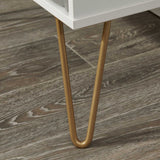 A white piece of furniture with slender, golden legs stands on a wooden floor with prominent, greyish grain patterns.